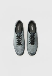 Grå lærersneakers med teksturert overflate, med sorte snører, avrundet tå og en subtil logo på innersålen.
