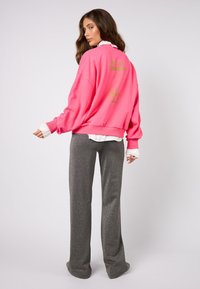 Nadrozmerná ružová mikina so zeleným textom, vysokým golierom a rebrovanými manžetami, doplnená o sivé široké nohavice so trblietavým efektom.