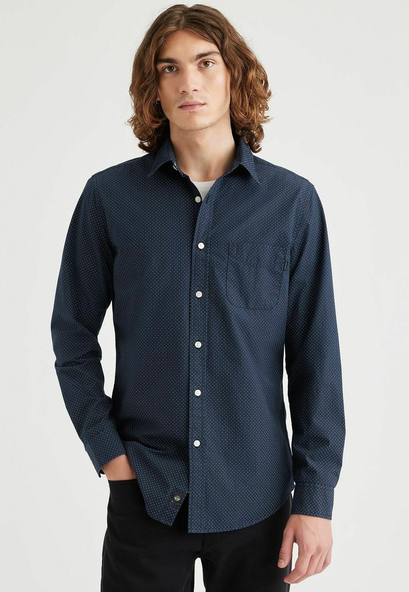 Marineblauwe langslavige shirt met een klein wit stippenpatroon, knoopsluiting aan de voorkant, enkele borstzak en een gebogen zoomontwerp.