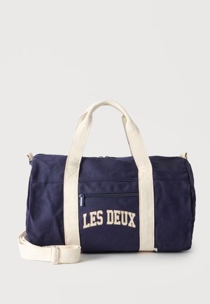 Sac polochon bleu marine avec poignées et sangle crème, poche zippée frontale et texte "LES DEUX" en lettres crème.