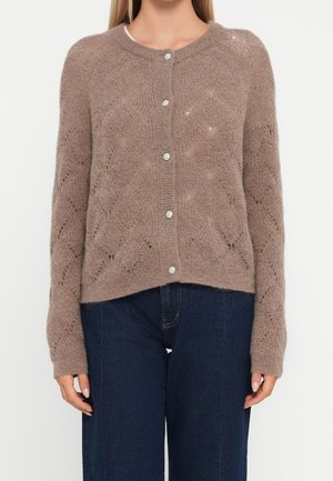 Kvinde iført en brun strikket cardigan med knapper og diamantmønstre samt blå cowboybukser, der står mod en ensfarvet hvid baggrund.