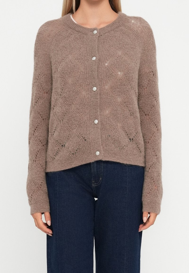 Femme portant un cardigan marron en tricot boutonné avec des motifs en losange et un jean bleu en denim, debout sur un fond blanc uni.