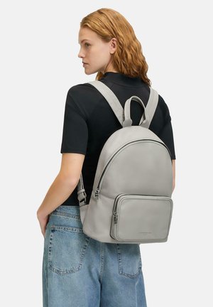 Grauer Leder-Rucksack mit runder Form, vorderer Reißverschlusstasche, verstellbaren Trägern und einem oberen Griff. Tragen auf dem Rücken vor einem schlichten Hintergrund.