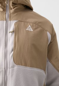 Tan og grå hybridjakke med glatt ytterstoff og myk fleece. Inkluderer et trekantet logo, glidelås og justerbar hette.