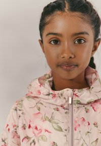 Veste rose clair à motif floral avec capuche, dotée d'une fermeture éclair à l'avant, texture du tissu lisse, agrémentée de feuilles vertes et de roses.