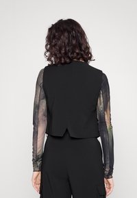Gilet nero aderente con un design strutturato, caratterizzato da una texture liscia e uno spacco sul retro, abbinato a maniche lunghe trasparenti e patterned in colori tenui.