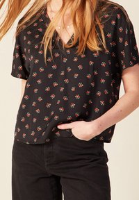 Donna che indossa una blusa nera a maniche corte con piccole fiori rossi e pantaloni neri, con una mano in tasca.
