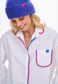 Biała koszula z różowymi i niebieskimi akcentami, z kieszenią z niebieską metką. Na głowie miękka fioletowa dziergana czapka.