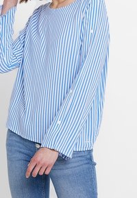 Osoba w niebiesko-białą pionową w paski koszulę z długim rękawem, połączoną z niebieskimi dżinsami, pozująca na jednolitym białym tle.