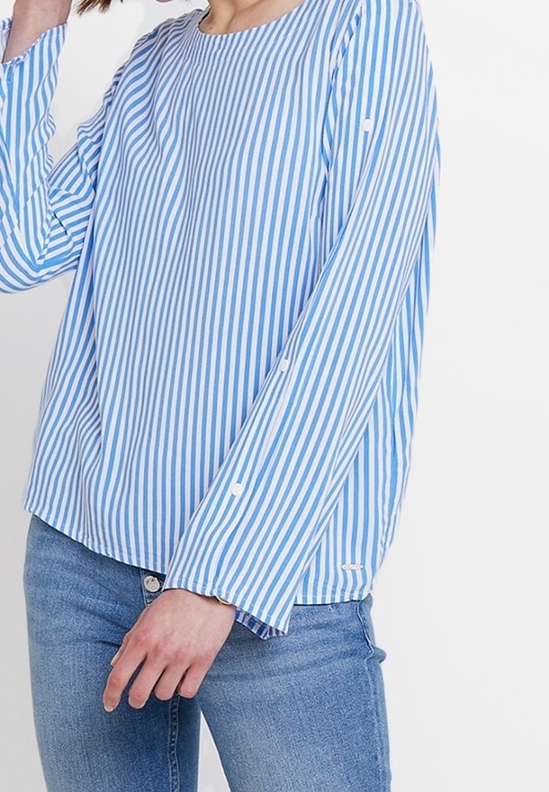 Osoba w niebiesko-białą pionową w paski koszulę z długim rękawem, połączoną z niebieskimi dżinsami, pozująca na jednolitym białym tle.