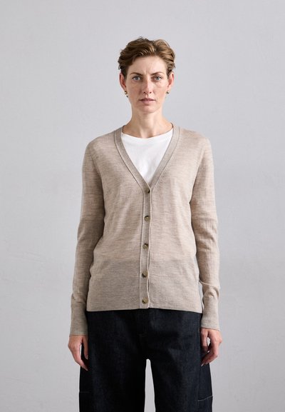 Beige Strickjacke mit V-Ausschnitt, Knopfleiste und zwei vorderen Taschen, kombiniert mit einem weißen T-Shirt und dunklen Hosen. Weiche Textur, taillierte Passform.