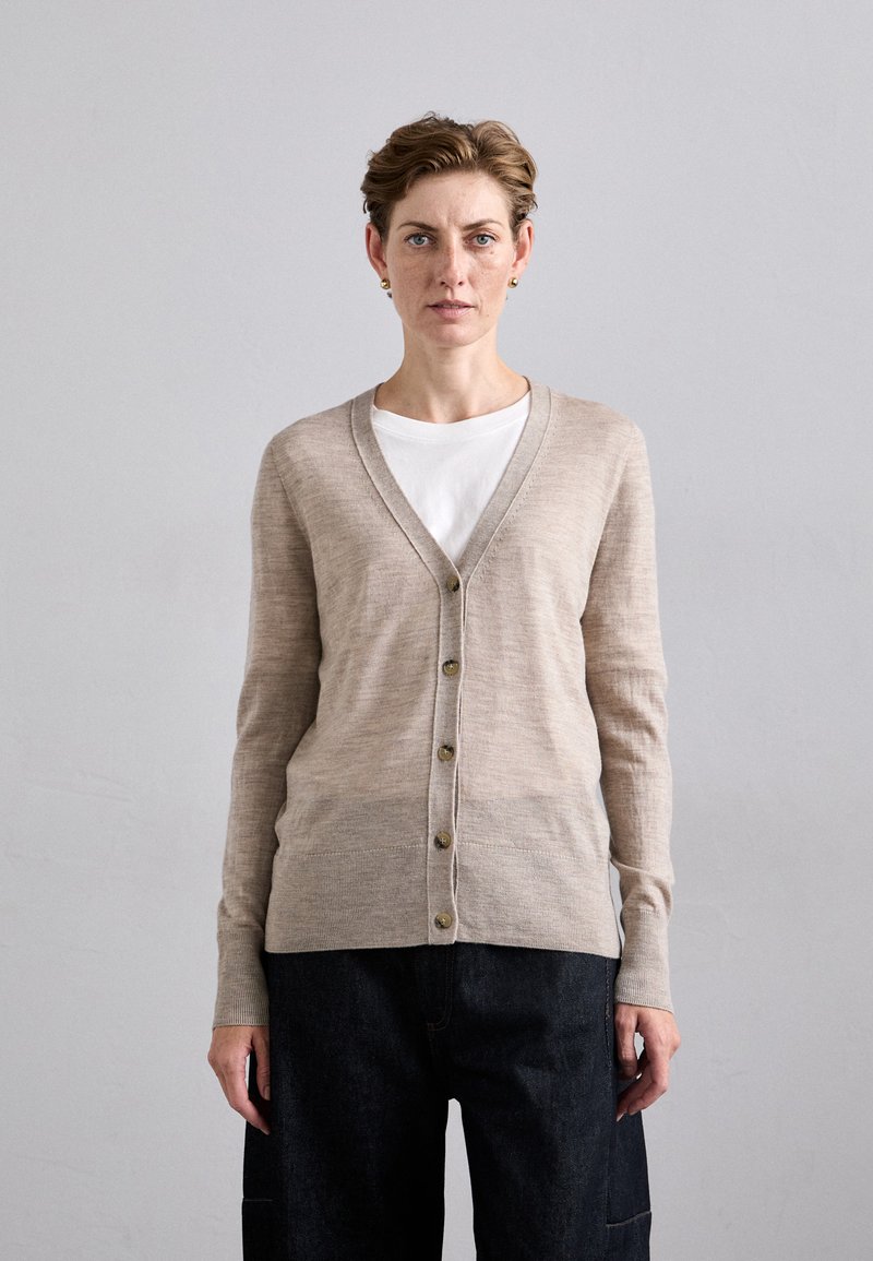 Beige Strickjacke mit V-Ausschnitt, Knopfleiste und zwei vorderen Taschen, kombiniert mit einem weißen T-Shirt und dunklen Hosen. Weiche Textur, taillierte Passform.