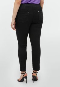 Jeans negros ajustados con una textura suave, que presentan dos bolsillos traseros y tachuelas en tono dorado, ajustados en el tobillo. Llevados con sandalias de tacón negras.