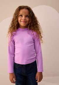 Langærmet ribstrikket top i lys lilla med rund halsudskæring, kombineret med mørkeblå jeans. Glat tekstur, tætsiddende design.