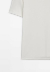 Une partie d'un T-shirt beige uni à manches courtes, montré sur un fond blanc.