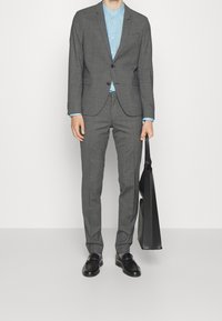 Completo grigio a due pezzi con taglio su misura, camicia azzurro chiaro e scarpe di pelle nere. Tiene una borsa nera; texture liscia e design strutturato.