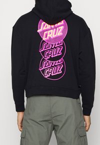 Svart croppad hoodie med tre stora cirkulära rosa tryck med texten "SANTA CRUZ", som visar en slät textur och huva.