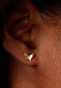 Boucles d'oreilles en or en forme de papillon avec des surfaces lisses et réfléchissantes, positionnées sur une oreille texturée près du lobe.