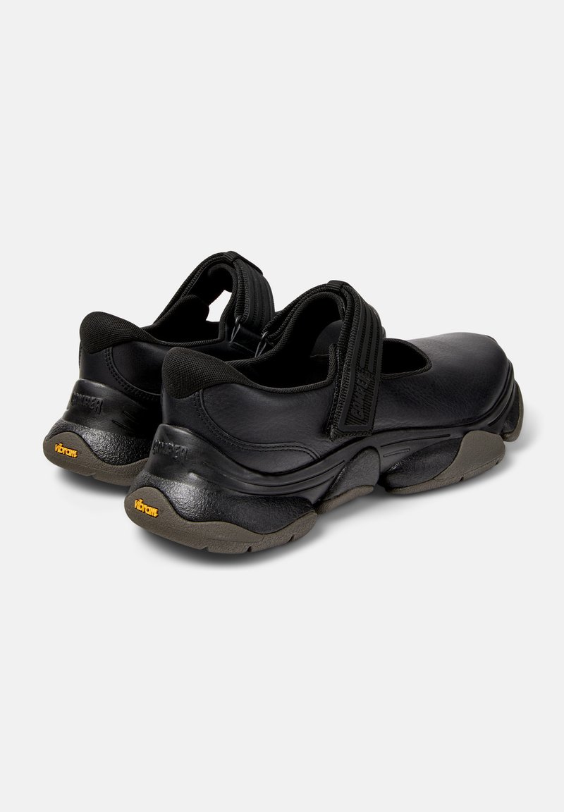 Une paire de baskets Mary Jane en cuir noir avec des semelles épaisses, des bandes Velcro et un marquage Vibram au talon et sur la semelle extérieure.