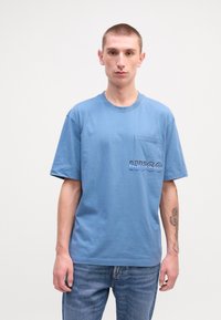 Modrá bavlnená tričko s krátkymi rukávmi, ktorá má náprsné vrecko a logo "HUGO" v kontrastných farbách. Nosené s svetlomodrým džínsy.