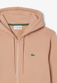 Sudadera con cremallera de color melocotón hecha de tela suave, con una capa interior verde, capucha con cordón y un pequeño logo de cocodrilo bordado.