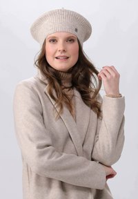 Beige wool blend beret with ribbed texture and small metal accent, paired with a light beige coat and a fitted turtleneck.