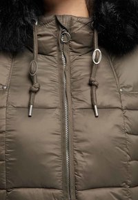 Veste rembourrée vert olive avec fermeture éclair, capuche à cordon de serrage et bordure en fausse fourrure noire. Comprend des accessoires argentés et des sections cousues.