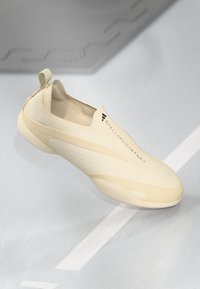 Beige instap sneaker met een minimalistisch ontwerp, subtiele texturen en "Stella McCartney" branding aan de bovenkant tegen een grijze achtergrond.