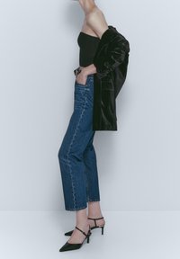 Haut noir sans bretelles, jean droit délavé clair, manteau en velours noir posé sur l'épaule, et chaussures à talons noires à bout pointu.