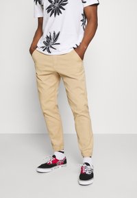 Beige chino's met elastische boorden, gecombineerd met een wit t-shirt met zwarte bloemenprint en zwart/witte geblokte Vans sneakers.