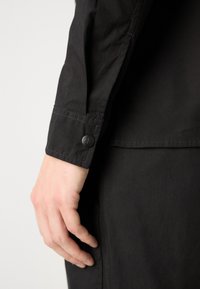 Μαύρο πουκάμισο με μακριά μανίκια, που διαθέτει κουμπωτό μανίκι, λεία υφή και διακριτή γυαλάδα, με ορατό λογότυπο της επωνυμίας στο κουμπί.