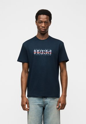 Tânăr purtând un tricou bleumarin cu inscripția "1985 Tommy Jeans" și blugi din denim bleu deschis, stând în fața unui fundal alb simplu.