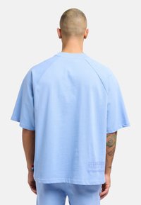 Light blue oversized t-shirt with short raglan sleeves, smooth fabric, and back text detail reading "Established Milano 1991."