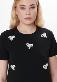 Schwarzes kurzärmliges T-Shirt mit sechs weißen, mit Perlen verzierten Schleifen, die an verschiedenen Stellen auf der Vorderseite angeordnet sind. Glatte Stofftextur.