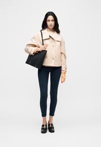 Γυναίκα που φοράει μπεζ παλτό τύπου pea coat, στενά μαύρα παντελόνια, μαύρα loafers με χρυσές αλυσίδες και κρατάει μαύρη τσάντα ώμου σε λευκό φόντο.