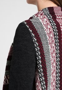 Virknad cardigan med en mönstrad vinröd och svart bakdel samt enfärgade svarta ärmar; har en texturerad look och kontrasterande färgschema.