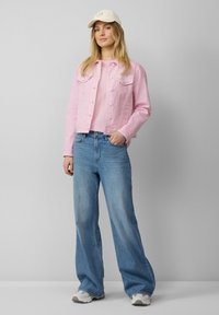 Roze denimjack met twee voorzakken, gedragen over een lichtroze shirt. Blauwe wijde jeans met een hoge taille en sneakers maken de outfit compleet.