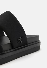 Crne sandale s širokim tkanim trakama i teksturiranim đonom. 'cK' logo je urezan sa strane. Glatki, minimalni dizajn.