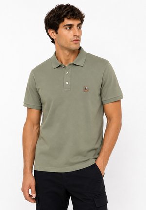 Homme portant un polo manches courtes vert olive avec un logo sur la poitrine et un pantalon noir, regardant vers sa droite avec la main dans la poche.