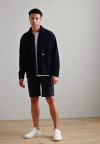 Ung mann står innendørs iført en svart jakke, hvit t-skjorte, svarte shorts og hvite joggesko på et tregulv.