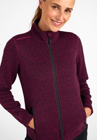 Mujer vistiendo una chaqueta de forro polar de rayas negras y marrón con cremallera, con las manos en los bolsillos, sonriendo contra un fondo blanco.