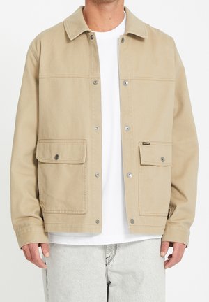 Mann trägt beige Jacke mit großen Fronttaschen über weißem Hemd und hellgrauen Jeans, stehend vor weißem Hintergrund.