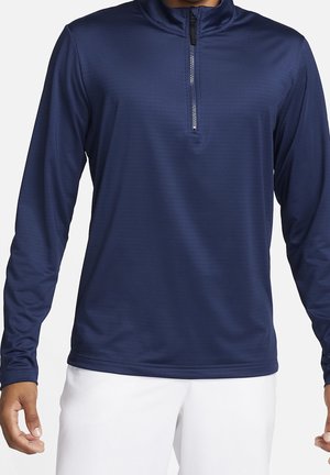 Mann trägt ein marineblaues, langärmeliges Sportshirt mit halbem Reißverschluss und weiße Hosen, steht mit entspannten Armen an den Seiten.