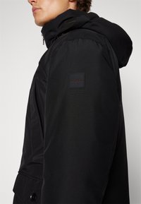 Homme portant une veste noire à capuche avec un patch logo BOSS sur le haut de la manche, montré de côté et incliné vers le haut.