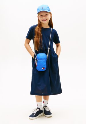 Niña con cabello largo y rojo, vestida con un vestido azul marino, gorra azul, calcetines blancos con logo, zapatillas azules marinas y un bolso cruzado azul, de pie con las manos en los bolsillos.
