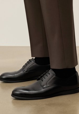 Czarne, skórzane, sznurowane buty wizytowe, noszone z ciemnymi skarpetkami i brązowymi, dopasowanymi spodniami, na tle neutralnej podłogi i ściany.