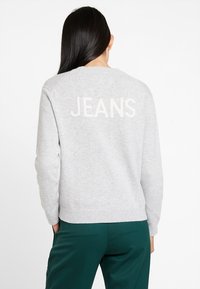 Grauer Pullover mit gerippten Bündchen und Saum, auf dessen Rückseite das Wort "JEANS" in Cremefarbe zu sehen ist. Glatte Textur, lockere Passform, kombiniert mit grünen Hosen.