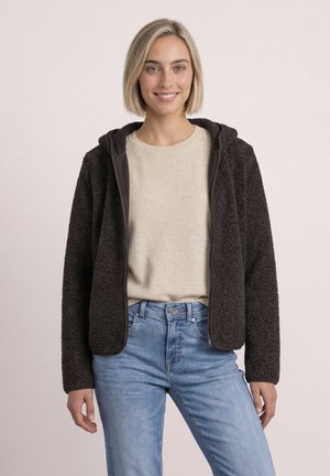 Jonge vrouw met kort blond haar, die een beige trui, een zwarte fleecejas met rits en lichtblauwe spijkerbroek draagt, glimlacht naar de camera.