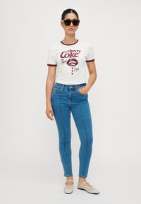 Λευκό μπλουζάκι με μπορντό γραφικό σχέδιο, συνδυασμένο με μπλέ στενά τζιν που διαθέτουν πλαϊνές ραφές, και μπεζ φλατ παπούτσια. Συμπληρωμένο με γυαλιά ηλίου.