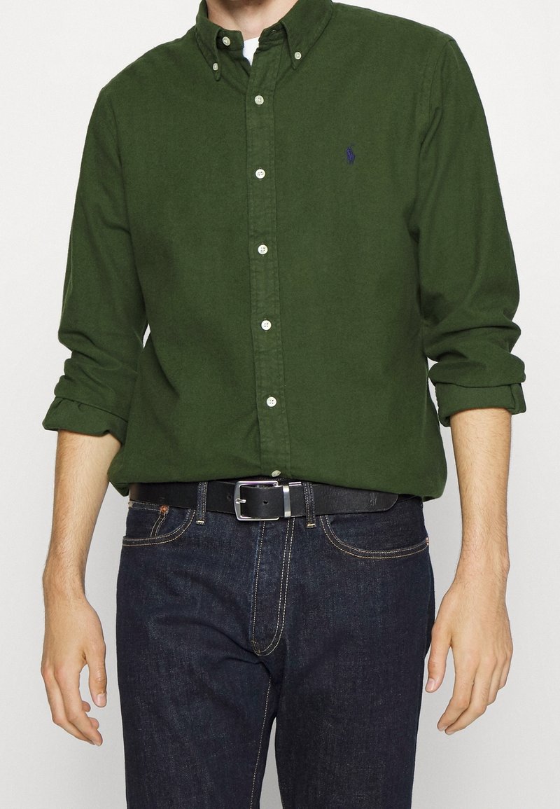 Homme portant une chemise boutonnée vert foncé avec les manches retroussées, un jean bleu foncé et une ceinture noire, debout devant un fond uni.